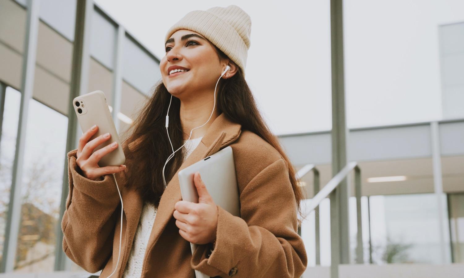 woman walking wearing headphones plugged in to a phone in one hand