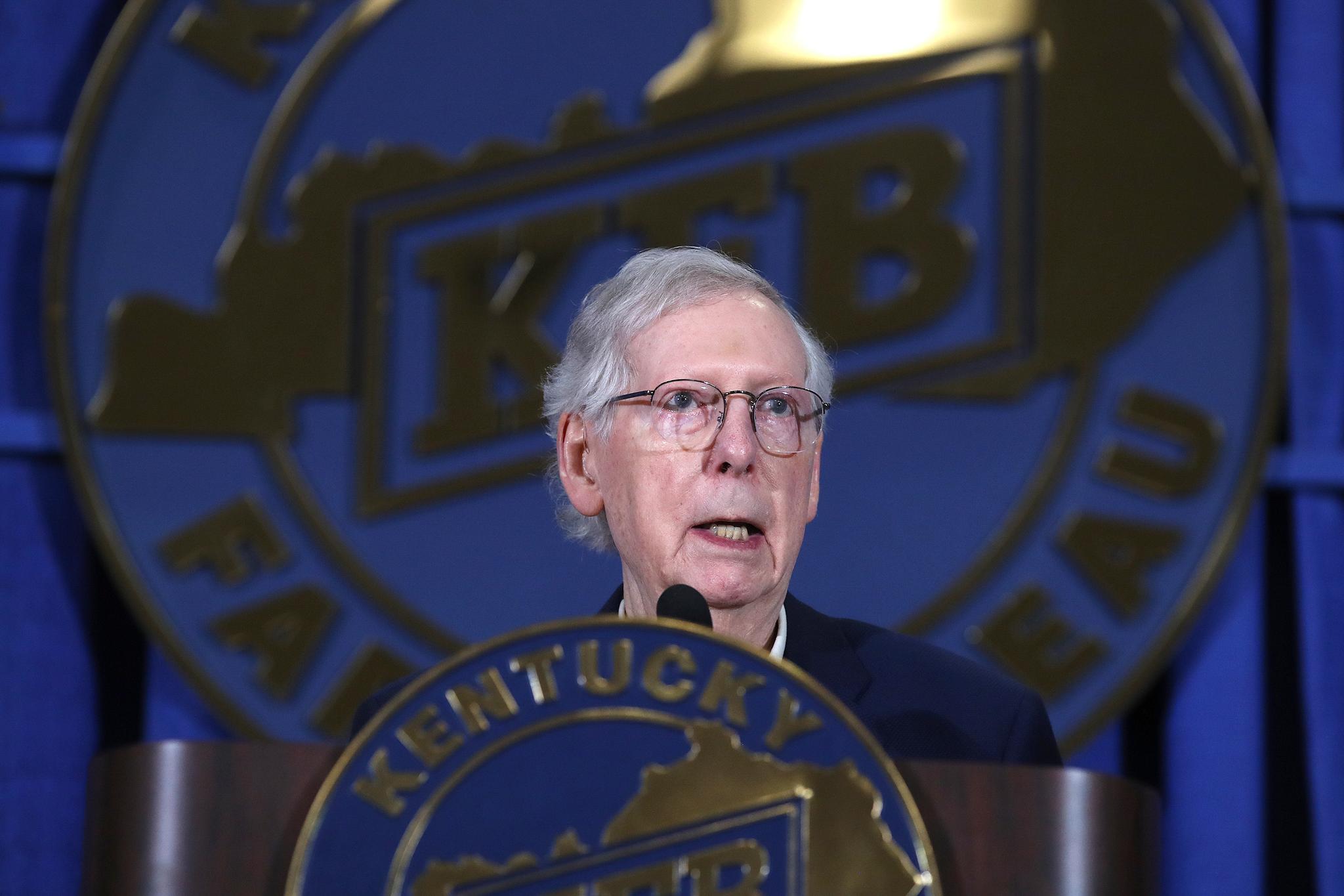 Mitch McConnell in front of a huge blue and gold emblem, speaking at a podium with a microphone