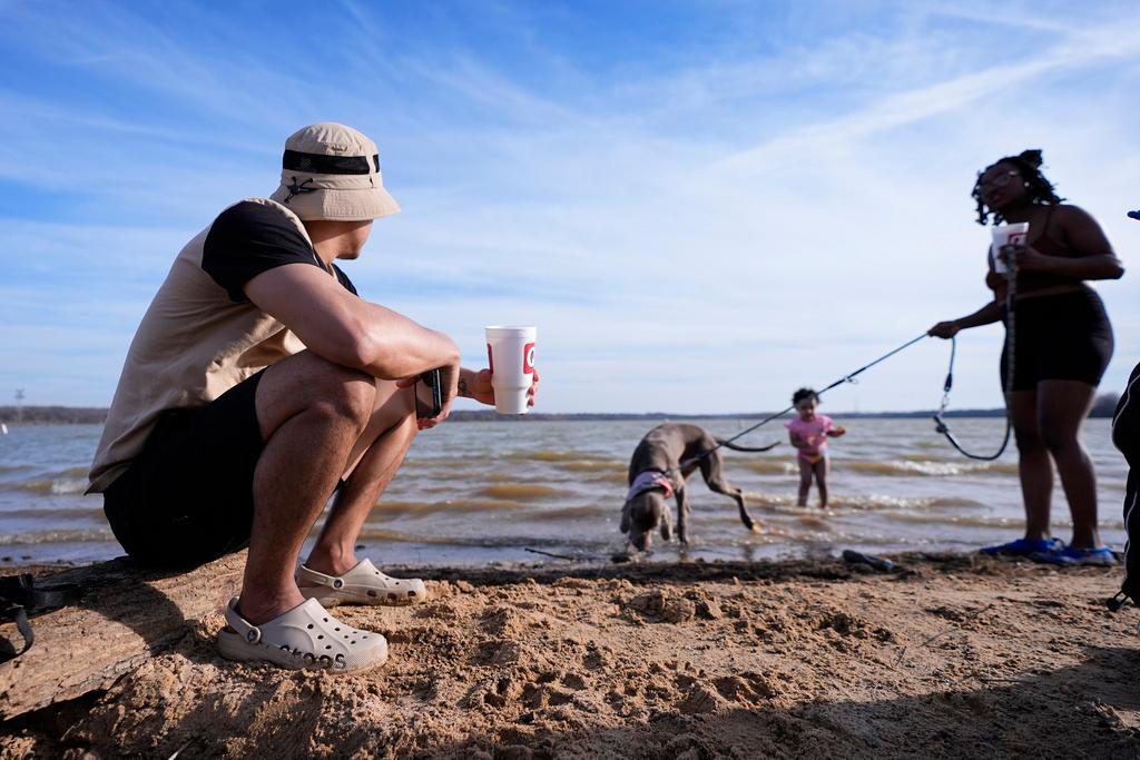 Victor Marcano, left, sits on a log as he watches his daughter Zhamira Marcano, 3, stand in the water on the shore of Joe Pool Lake with Joanna Clarkley, right, and her dog, Pluto, during an unseasonably warm winter day in Grand Prairie, Texas