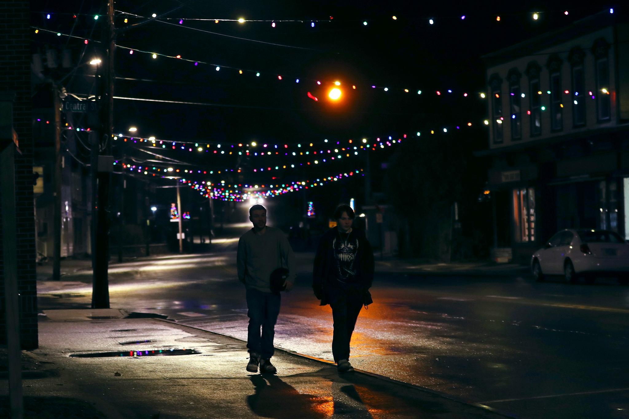 In this March 19, 2020, photo, two young men walk down Main Street under Christmas lights through downtown in Farmington, N.H. As the coronavirus spreads, holiday lights are going back up to provide a bit of emotional and actual brightness. 